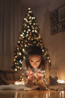 Güzel genç bir kadın güzel dekore edilmiş Noel ağacının yanında yerde yatıyor evde dinleniyor ve elinde Noel ışıklarıyla kış mevsiminin tadını çıkarıyor.