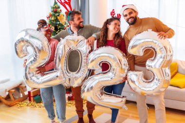 Bir grup genç arkadaş, 2023 numaralı dev balonları tutarak evde yeni yılı kutluyor.