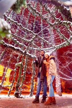 Güzel genç çift Noel pazarında eğleniyor, dekore edilmiş şehir sokaklarında yürüyor, sıcak sıcak şarap içiyor ve sarılıyorlar.