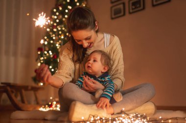Anne bebeğini kucağında tutuyor, Noel arifesini evde kutluyor, güzel dekore edilmiş Noel ağacının yanında oturuyor, elinde maytap tutuyor.
