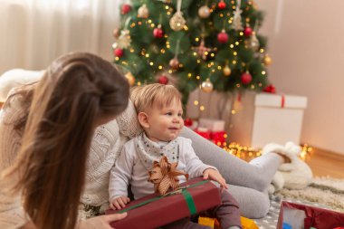 Anne ve bebek Noel günü evde eğleniyor, güzel dekore edilmiş Noel ağacının yanında oturuyor, hediyeleri açıyor ve oyuncaklarla, yaylarla ve kurdelelerle oynuyor.