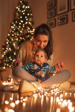 Anne bebeğini kucağında tutuyor, güzel süslenmiş Noel ağacının yanında oturuyor, Noel arifesini evde kutluyor.