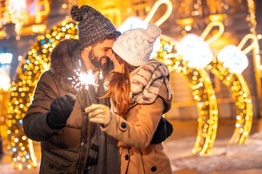 Aşıklar şehir meydanında yeni yılı kutlarken ellerinde gece yarısı geri sayımı için maytap tutarken ve Noel ışıkları yanarken eğleniyorlar.