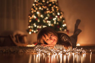Güzel küçük kız Noel 'i evde kutlarken eğleniyor, yere uzanıyor, güzelce süslenmiş Noel ağacının yanında ve gülümsüyor.