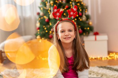 Geyik boynuzu takan güzel bir kız Noel ağacının yanında oturmuş Noel 'i evde kutlarken eğleniyor.