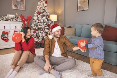 Genç ebeveynler bebekleriyle oynarken Noel 'i evde birlikte geçirip, balon üflerken ve fırlatırken eğleniyorlar.