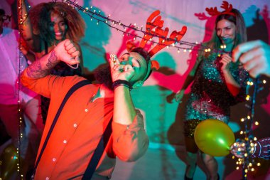 Bir grup neşeli genç arkadaş yılbaşı partisinde yılbaşı ışıklarının altında dans ederek eğleniyorlar.