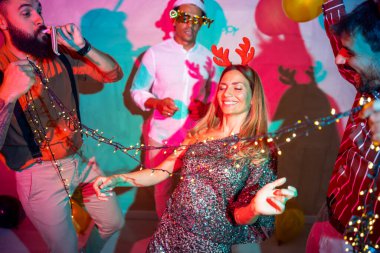 Bir grup neşeli genç arkadaş yılbaşı partisinde yılbaşı ışıklarının altında dans ederek eğleniyorlar.