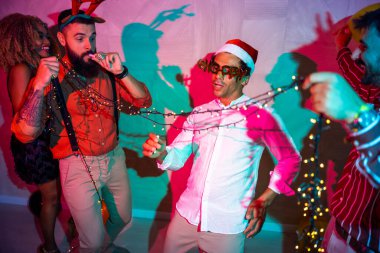 Bir grup neşeli genç arkadaş yılbaşı partisinde yılbaşı ışıklarının altında dans ederek eğleniyorlar.