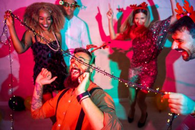 Bir grup neşeli genç arkadaş yılbaşı partisinde yılbaşı ışıklarının altında dans ederek eğleniyorlar.