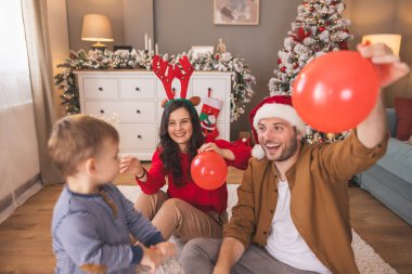 Genç ebeveynler bebekleriyle oynarken Noel 'i evde birlikte geçirip, balon üflerken ve fırlatırken eğleniyorlar.