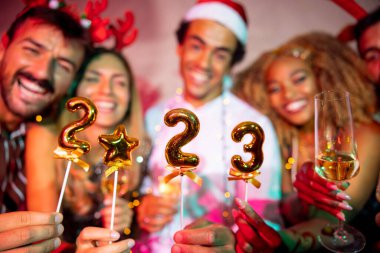 Bir grup neşeli genç arkadaş yeni yıl partisinde eğleniyor, gece yarısı geri sayımında şarkı söylüyor ve dans ediyor, ellerinde 2023 numaralı balonlar var.