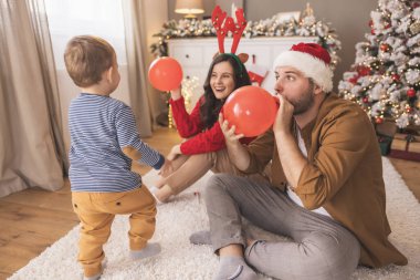 Genç ebeveynler bebekleriyle oynarken Noel 'i evde birlikte geçirip, balon üflerken ve fırlatırken eğleniyorlar.