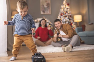 Genç ebeveynler, Noel 'i evde birlikte geçirirken küçük çocuklarıyla oynarken, uzaktan kumandalı oyuncak arabaları kovalarken eğleniyorlar.