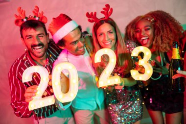 Yeni yıl partisinde eğlenen bir grup arkadaş, 2023 yılını gece yarısı geri sayımını temsil eden aydınlatıcı numaralar tutuyorlar. 20 numaraya odaklan.