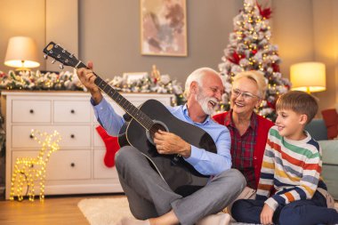 Küçük çocuk kış tatilini büyükannesi ve büyükbabasıyla geçirirken, büyükbabası gitar çalarken karısı ve torunu Noel günü evde şarkı söylerken eğleniyor.