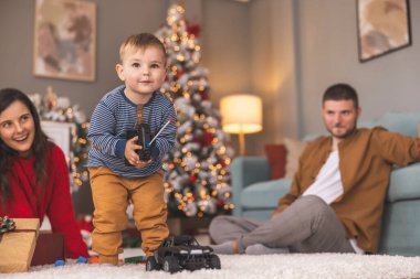 Genç ebeveynler Noel 'i evde birlikte geçirirken bebekleriyle oynarken eğleniyorlar, çocuk uzaktan kumandalı oyuncak arabalarla oynamayı öğreniyor.