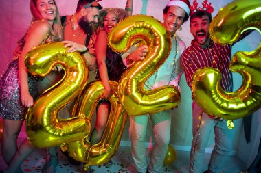 Bir grup genç arkadaş yeni yıl partisinde eğleniyor, dans ediyor ve gece yarısı geri sayımında 2023 numara şeklinde balonlar tutuyor.