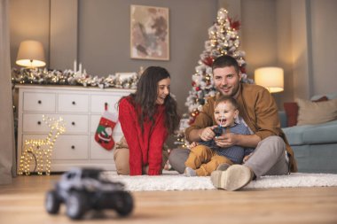 Genç ebeveynler, Noel 'i evde birlikte geçirirken çocuklarıyla oynarken eğleniyorlar. Baba, oğluna uzaktan kumandalı oyuncak araba kullanmayı öğretiyor.