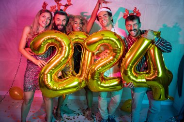 Bir grup genç arkadaş yeni yıl partisinde eğleniyor, dans ediyor ve gece yarısı geri sayımında 2024 numara şeklinde balonlar tutuyor.