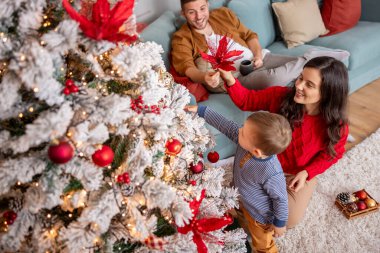 Genç ebeveynler, Noel ağacını şirin erkek bebekleriyle süsleyerek, Noel süsleri takıp, kış tatili için evi dekore ederek eğleniyorlar.