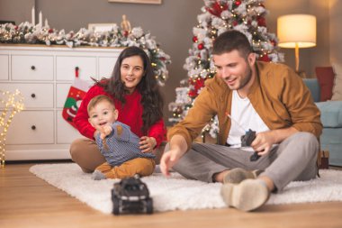 Genç ebeveynler Noel 'i evde birlikte geçirirken bebekleriyle oynarken eğleniyorlar, çocuk uzaktan kumandalı oyuncak arabalarla oynamayı öğreniyor.