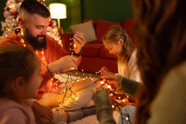 Ebeveynler ve çocuklar yılbaşı gecesini evde birlikte geçiriyorlar, Noel ağacının yanında oturuyorlar, Noel ışıklarıyla oynuyorlar.