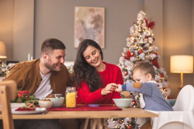 Genç ebeveynler yeni yürümeye başlayan çocuklarıyla Noel sabahı kahvaltı ediyorlar, bebek yüksek sandalyede oturup kaşık tutuyor ve yemekleri karıştırıyor.