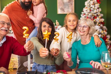 Mutlu çok nesilli aile, kış tatilini birlikte geçirmenin keyfini çıkarıyor, 2024 numaralı balonları tutarken, evde Noel yemeği yerken eğleniyor.
