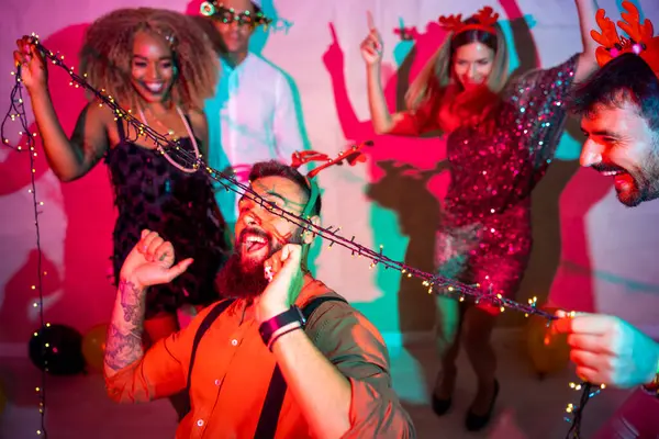 Bir grup neşeli genç arkadaş yılbaşı partisinde yılbaşı ışıklarının altında dans ederek eğleniyorlar.