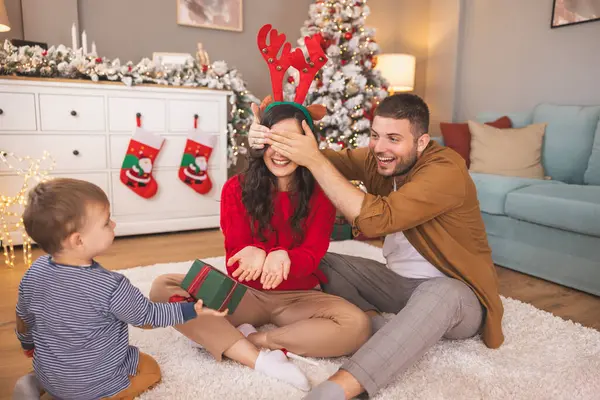 Genç ebeveynler Noel 'i evde bebekleriyle geçiriyorlar. Babaları annelerinin gözlerini kaparken, çocuklar ona Noel hediyesi veriyor.