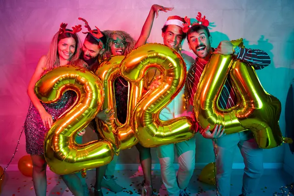 Bir grup genç arkadaş yeni yıl partisinde eğleniyor, dans ediyor ve gece yarısı geri sayımında 2024 numara şeklinde balonlar tutuyor.