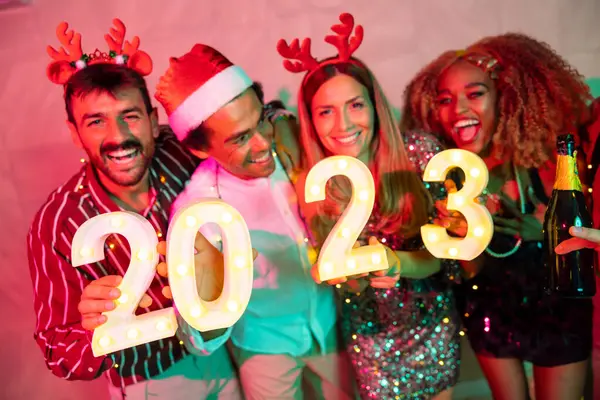 Yeni yıl partisinde eğlenen bir grup arkadaş, 2023 yılını gece yarısı geri sayımını temsil eden aydınlatıcı numaralar tutuyorlar. 20 numaraya odaklan.