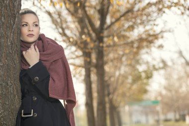 Güzel genç sarışın bir sonbahar günü bir ağaca yaslanıyor, dalgın mesafeye bakıyor.