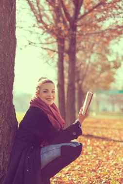 Güzel genç sarışın bir sonbahar günü bir ağaca yaslanıp kitap okuyor.