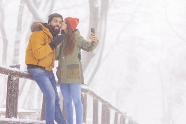 Güzel, genç bir çift, kış günü dışarıda dinlenirken ve sıcak çay içerken akıllı telefonlar kullanarak selfie çekerek eğleniyor.