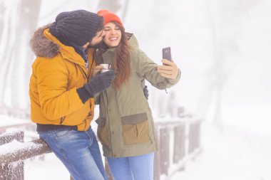 Güzel, genç bir çift, kış günü dışarıda dinlenirken ve sıcak çay içerken akıllı telefonlar kullanarak selfie çekerek eğleniyor.