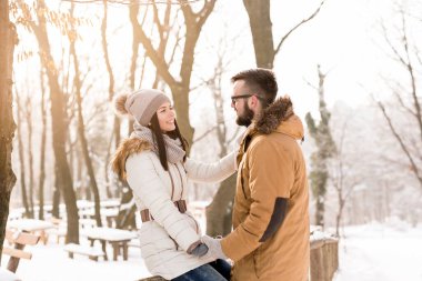 Birbirine aşık bir çift ormanda ahşap bir çitin yanında duruyor, konuşuyor ve güneşli bir kış gününün tadını çıkarıyor.
