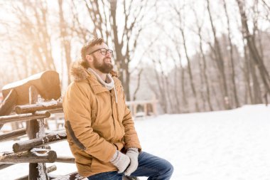Sıcak kışlık kıyafetler giyen yakışıklı genç adam, tahta çitlere yaslanıyor ve karlı bir kış gününün tadını çıkarıyor.