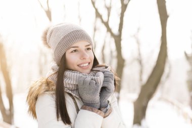 Güzel genç bir kadın ayakta, atkısını tutuyor ve gülümsüyor, güzel bir kış gününün tadını çıkarıyor.