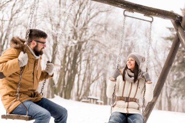 Beautiful young couple sitting on swings and having fun on a snowy winter day clipart