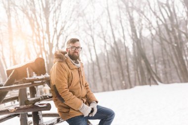 Sıcak kışlık kıyafetler giyen yakışıklı genç adam, tahta çitlere yaslanıyor ve karlı bir kış gününün tadını çıkarıyor.