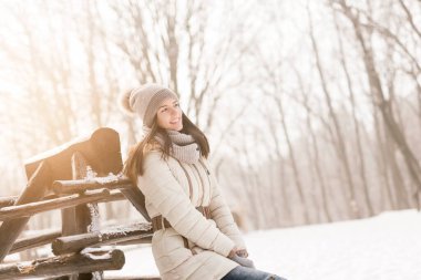 Sıcak kışlık kıyafetler giyen çekici genç bir kadın, ahşap bir heykele yaslanıyor ve karlı bir kış gününün tadını çıkarıyor.