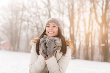 Güzel genç bir kadın ayakta, atkısını tutuyor ve gülümsüyor, güzel bir kış gününün tadını çıkarıyor.