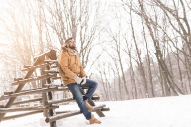 Sıcak kışlık kıyafetler giyen yakışıklı genç adam, tahta çitlere yaslanıyor ve karlı bir kış gününün tadını çıkarıyor.