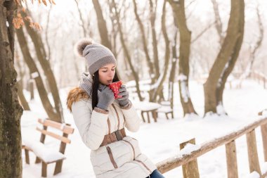 Güzel, genç bir kadın kışlık kıyafetler giyiyor, dışarıda duruyor ve bir fincan sıcak çay içiyor, karlı kış gününün tadını çıkarıyor.