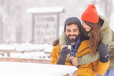 Güzel, genç ve aşık bir çift, karlı kış gününde ormanda yürüyüş yaparken, ahşap bankta oturur, sıcak çay içer, sarılır ve dinlenir.