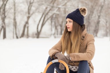 Güzel, genç bir kadın sıcak bir kış elbisesi giyiyor ve karlı bir kış gününde bir hünerin üzerinde oturuyor.