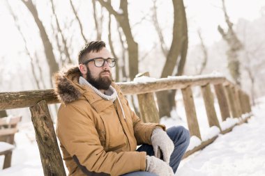 Sıcak kışlık kıyafetler giyen yakışıklı genç adam, tahta bir çite yaslanıyor ve dağlarda karlı bir kış gününün tadını çıkarıyor.