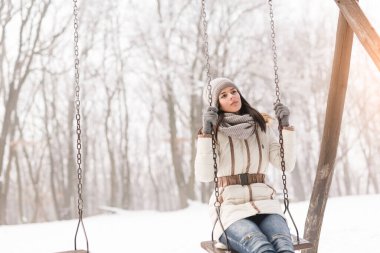 Güzel genç kız salıncakta oturuyor ve karlı bir kış gününün tadını çıkarıyor.